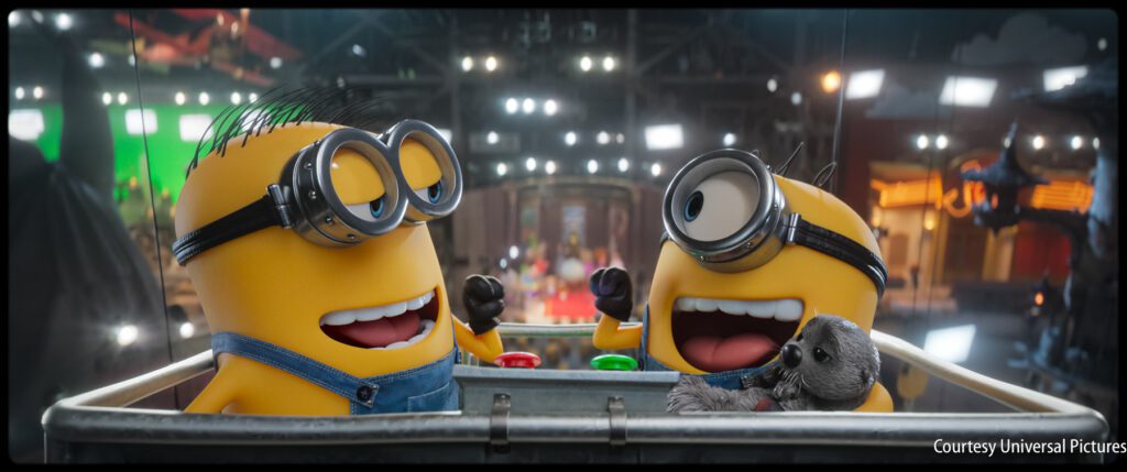Two animated yellow Minions in denim overalls laugh and gesture while sitting in a metal bin; one holds a stuffed animal, with bright lights and a blurred background.