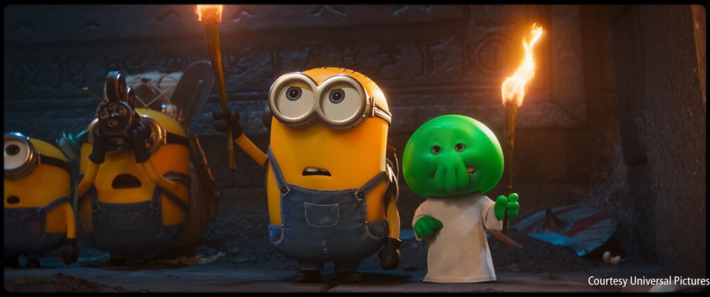 Three Minions holding torches stand beside a small green creature, all facing forward in a dimly lit stone chamber.