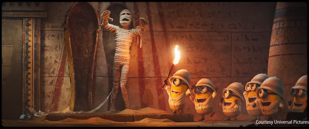 Six Minions in explorer hats look excitedly at a mummy emerging from a sarcophagus in an ancient, sand-filled Egyptian tomb.
