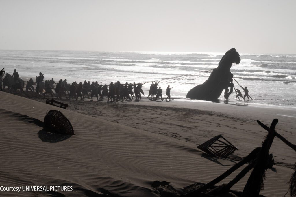 A group of people pull a large dinosaur sculpture with ropes along a sandy beach toward the ocean, with scattered debris in the foreground.