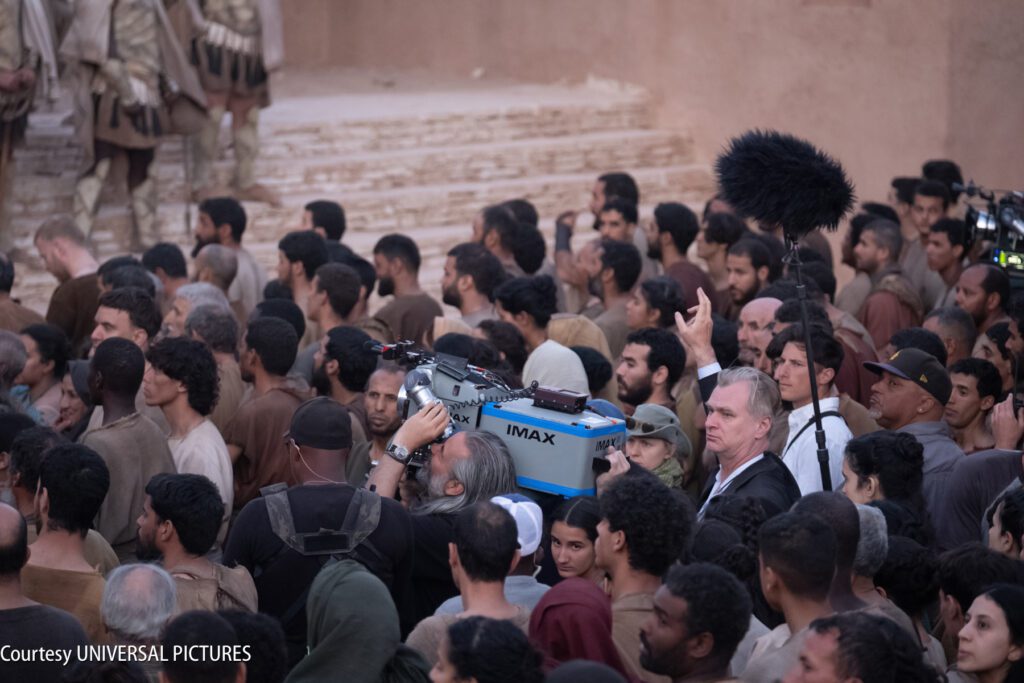 A film crew with an IMAX camera and boom microphone works among a large crowd of actors dressed in earth-toned costumes.