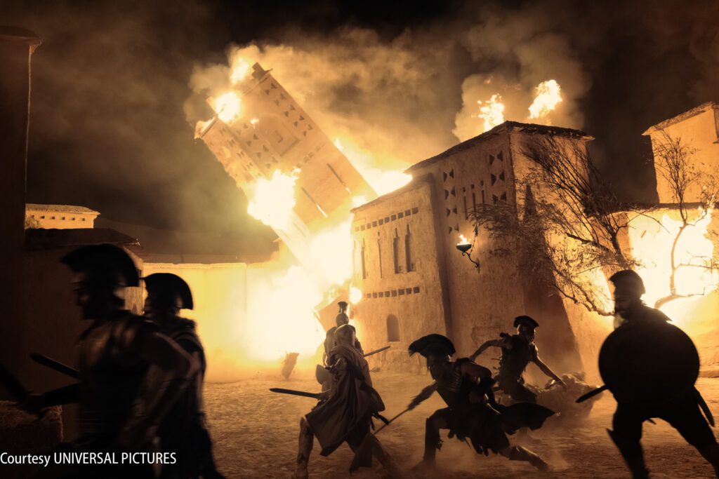 A group of armored soldiers run as a tall building collapses and flames engulf surrounding structures at night.