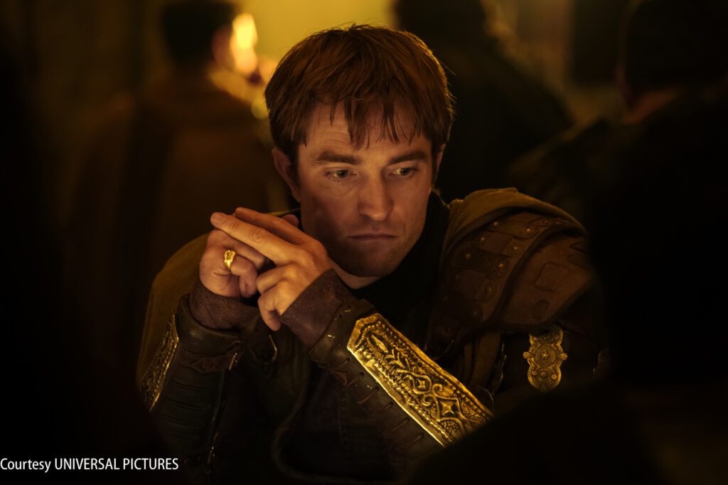 A man in medieval armor sits at a table with his hands clasped, looking thoughtful. Warm, dim lighting surrounds him.