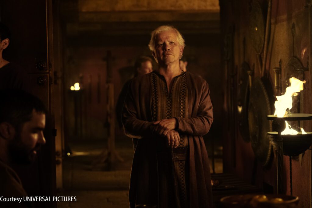 An older man in medieval clothing stands in a dimly lit room with torches and shields on the wall. Other people are visible in the background.