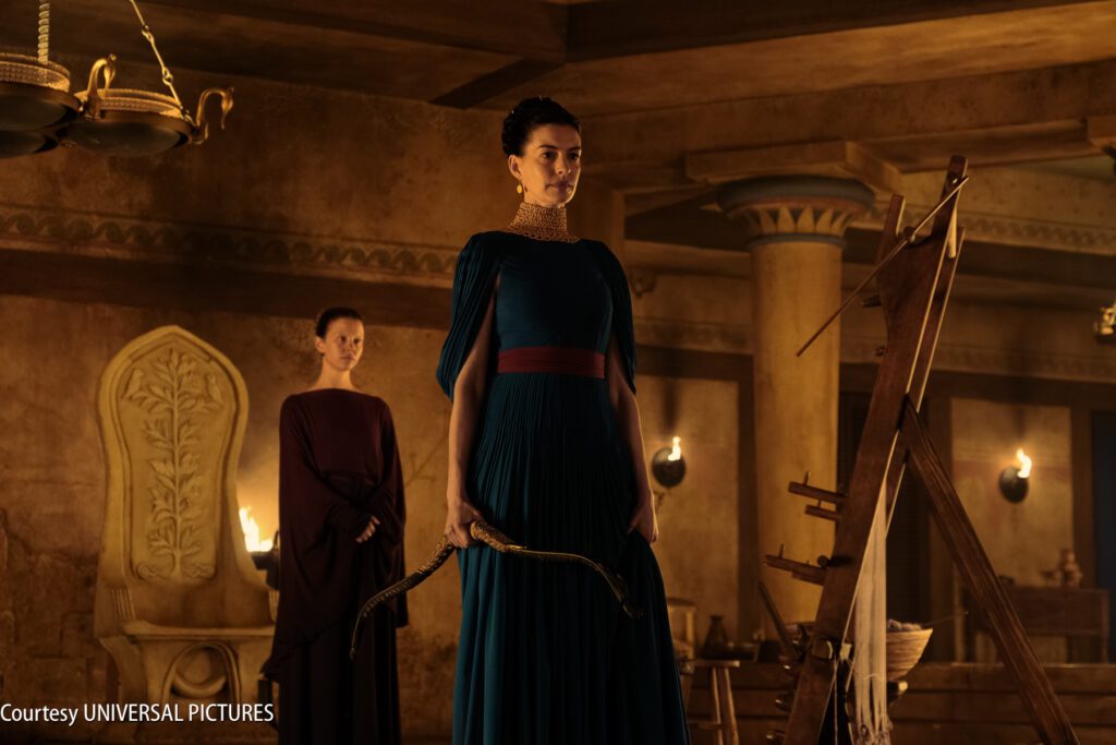 Two women in ancient-style clothing stand in a dimly lit, ornate room; one holds a bow next to an archery target, the other stands in the background.