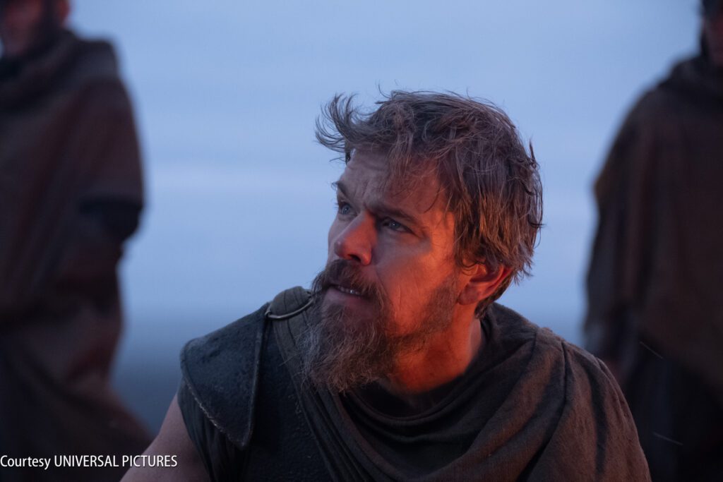 A bearded man in rugged clothing looks upward with a serious expression, against a blurred outdoor background at dusk.
