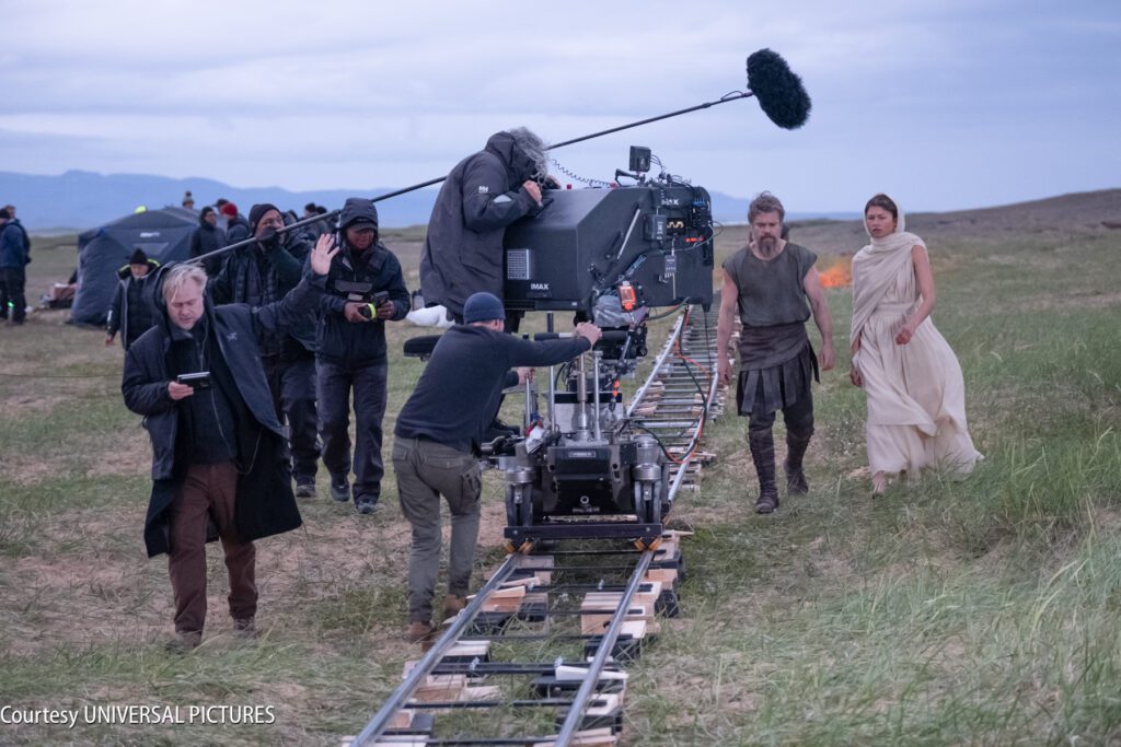 Film crew operates a camera on a track while two actors in costume walk nearby on a grassy field. Other crew members and equipment are visible in the background.