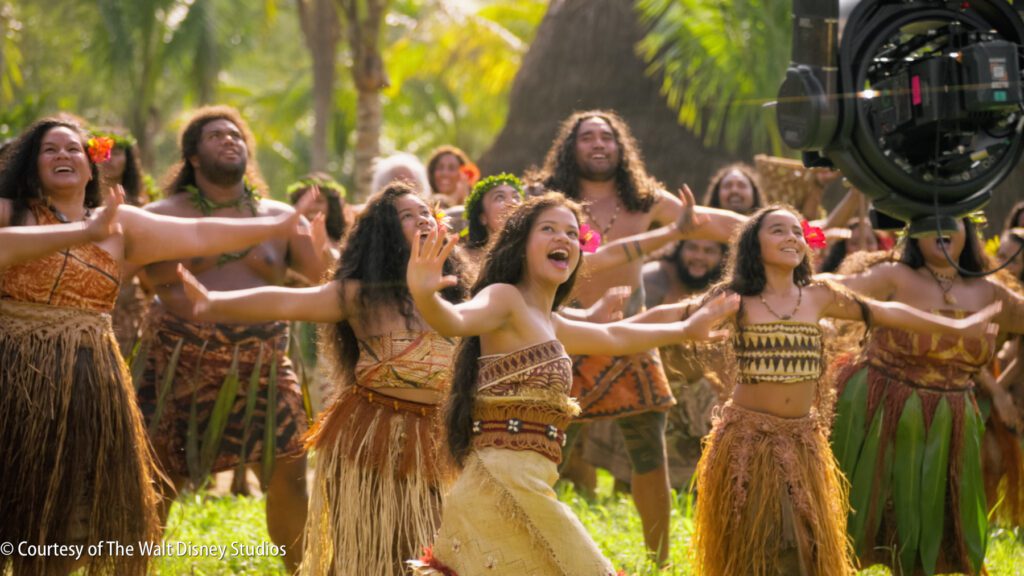 A group of people in Polynesian-inspired costumes perform a joyful dance outdoors, with a film camera visible on the right.