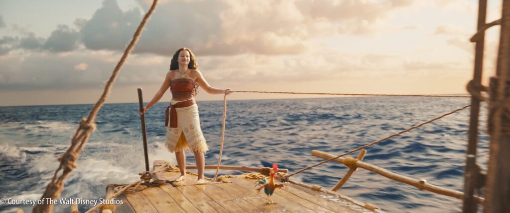 A young woman stands confidently on a wooden boat at sea, holding ropes, with a small rooster nearby. The sky is partly cloudy, and the ocean appears calm.