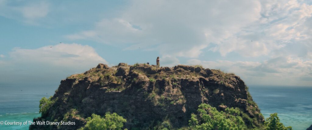A person stands alone on top of a rocky hill overlooking the ocean under a partly cloudy sky.