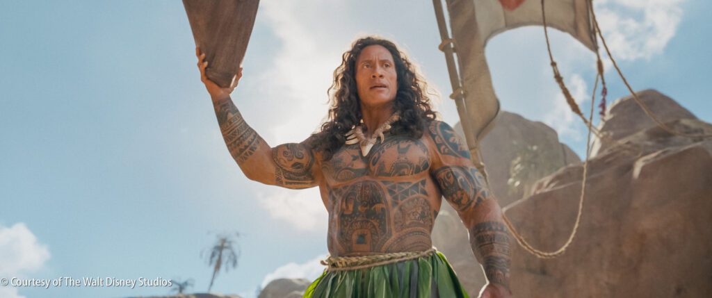 A muscular, tattooed man in a green skirt stands outdoors holding a wooden paddle, with rocks and blue sky in the background.