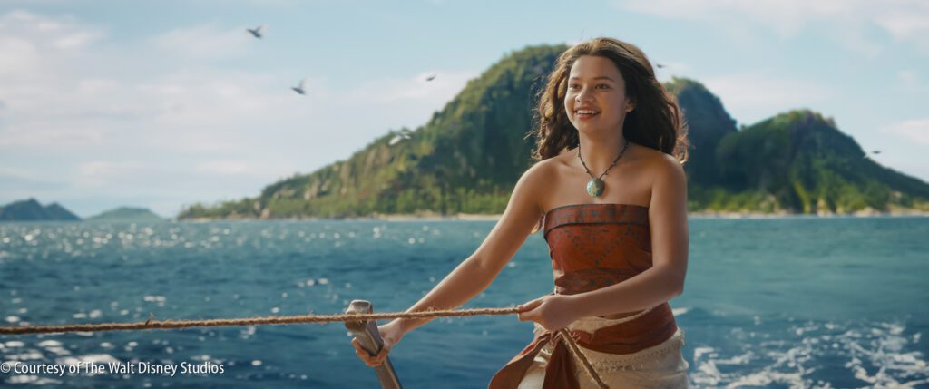 A young woman in island-inspired clothing stands on a boat holding a rope, with the ocean and a green island in the background under a blue sky.