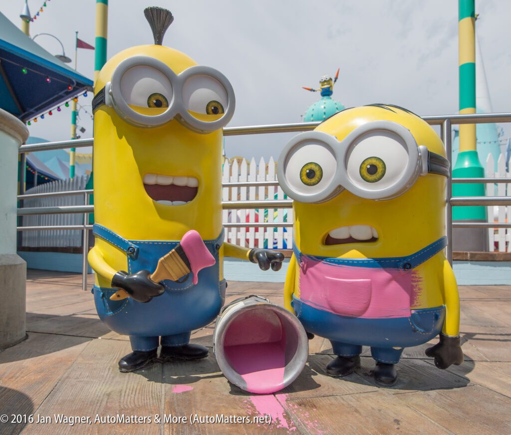Two Minion statues stand by a spilled can of pink paint on a wooden deck, one holding a paintbrush and smiling, the other looking surprised. Colorful amusement park structures are in the background.