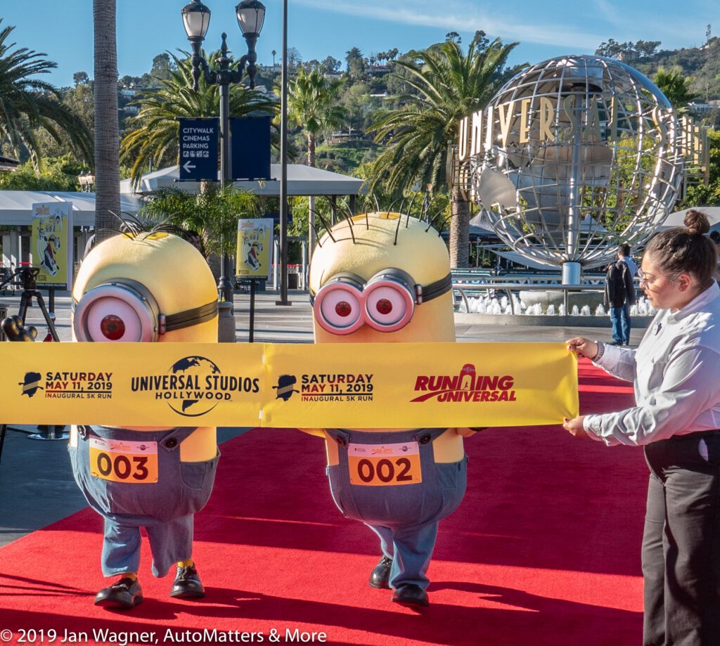 Two people in Minion costumes cross a finish line at Universal Studios Hollywood, with a staff member holding the banner and the Universal globe visible in the background.