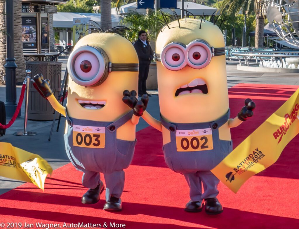 Two people in Minion costumes walk on a red carpet outdoors, wearing numbered bibs 003 and 002, holding yellow flags, with palm trees and event signs in the background.