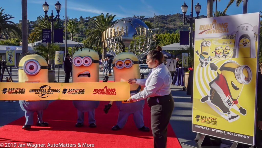 Three people in Minion costumes stand on a red carpet as a woman prepares to cut a ribbon for the Running Universal event at Universal Studios Hollywood.