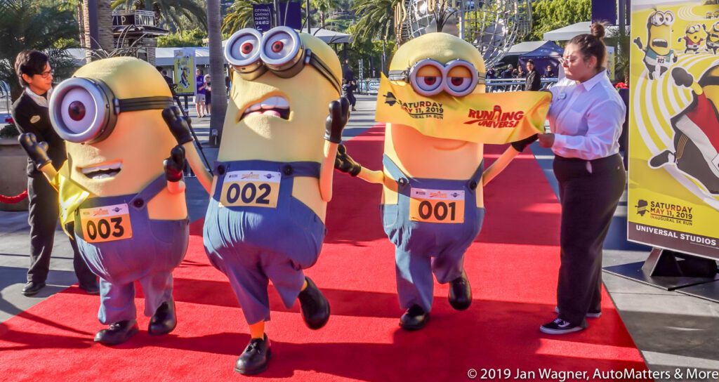 Three people in Minion costumes cross a finish line on a red carpet at an outdoor event, with a woman holding a yellow banner.