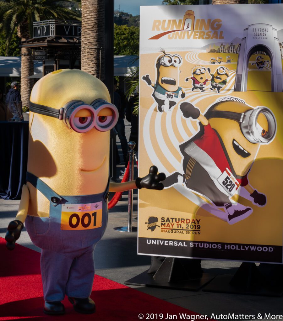 A person in a Minion costume with a race bib poses near a Running Universal 5K event sign at Universal Studios Hollywood.