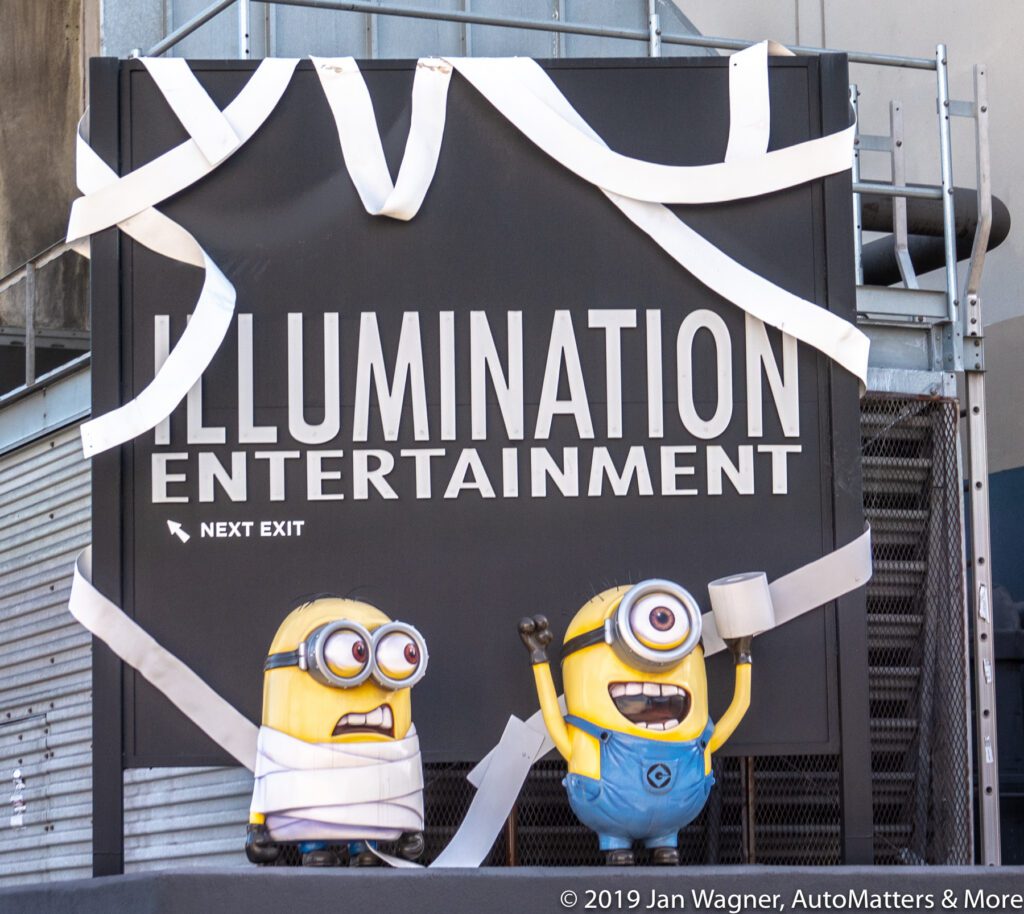 Two Minion statues, one holding a mug, stand in front of an "Illumination Entertainment" sign decorated with white streamers.