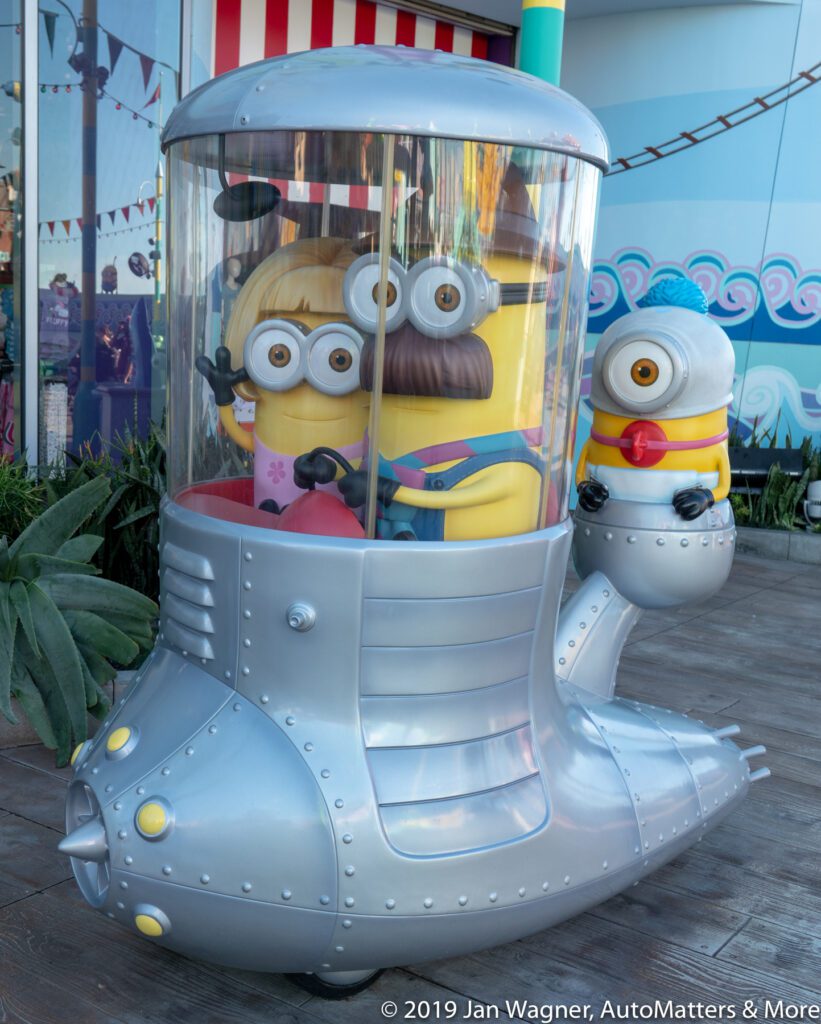 A group of Minion characters are inside a metallic, submarine-shaped amusement ride, with one Minion outside on a mechanical arm.