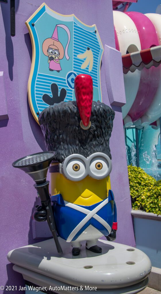 A statue of a Minion dressed as a royal guard stands against a purple wall with a decorative shield above it. The Minion holds a black staff and wears a tall black hat with a red plume.