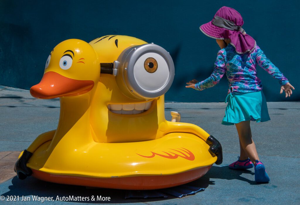 Young child in a sunhat approaches a yellow ride shaped like a hybrid duck and Minion character at an outdoor play area.