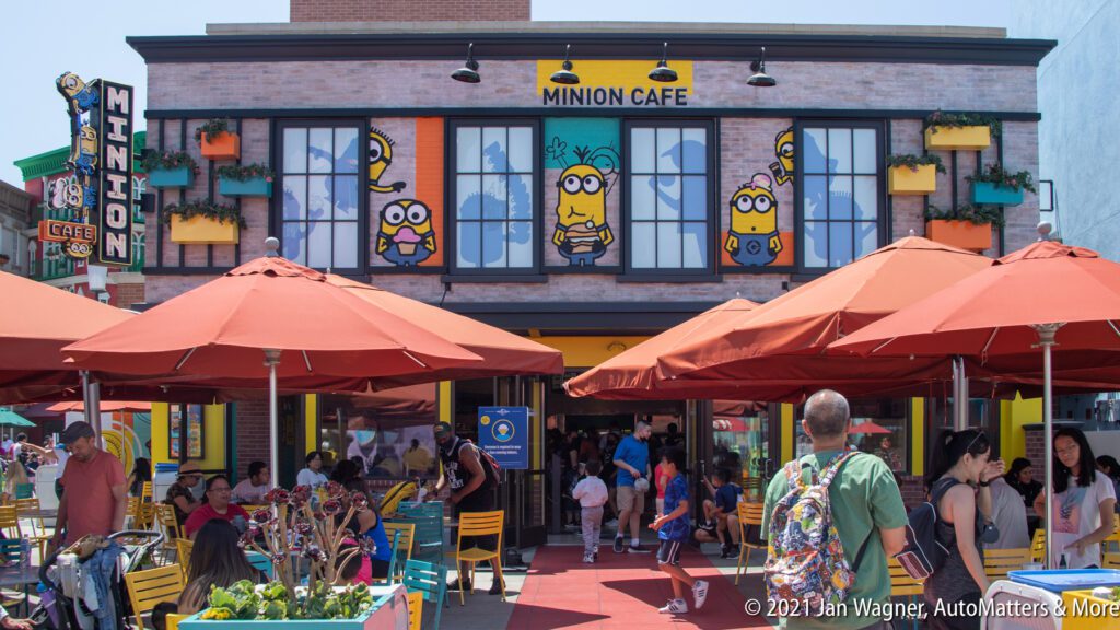 People walk near outdoor seating at the Minion Cafe, a themed restaurant with Minion decorations on the building, red umbrellas, and colorful plants.