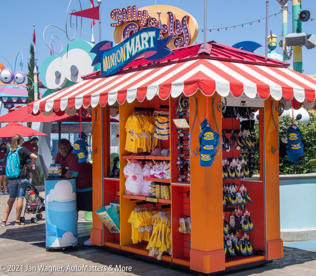 An orange and red-striped kiosk sells Minion merchandise, including clothing and toys, at an outdoor shopping area with people nearby.