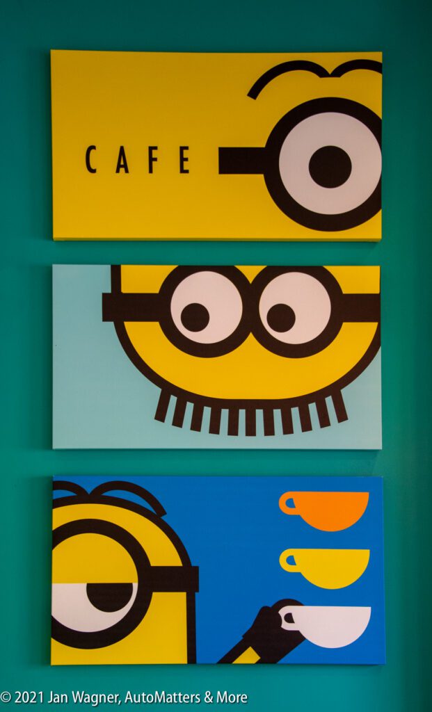 Three rectangular wall art pieces depict stylized cartoon yellow characters with goggles, referencing Minions, with coffee cups and the word "CAFE" on a teal background.