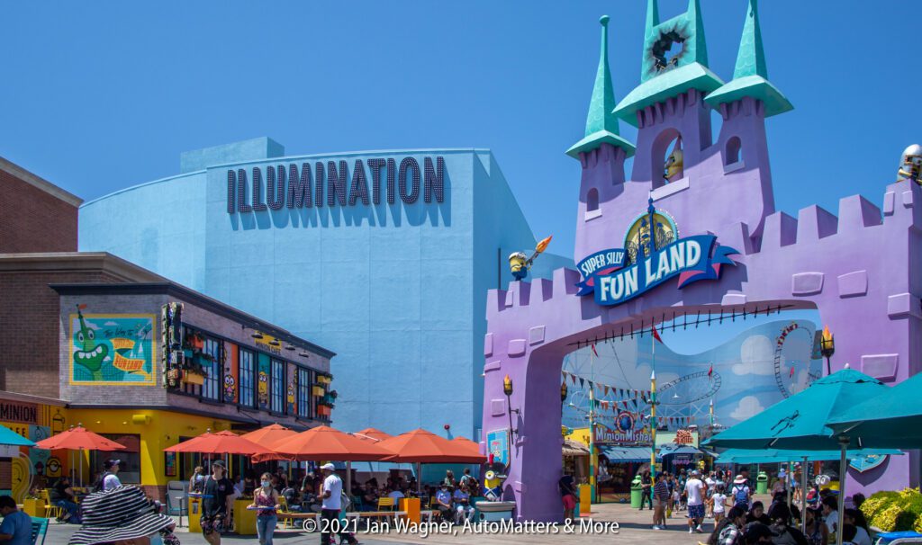 Crowds walk under colorful umbrellas near the entrance to Super Silly Fun Land, with the Illumination building visible in the background on a sunny day.