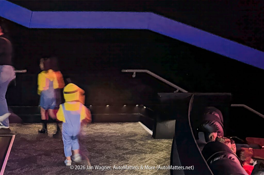 Two children in yellow Minion costumes walk up theater stairs, while others sit in seats and a third person walks ahead. The theater is dimly lit with black walls and blue accents.