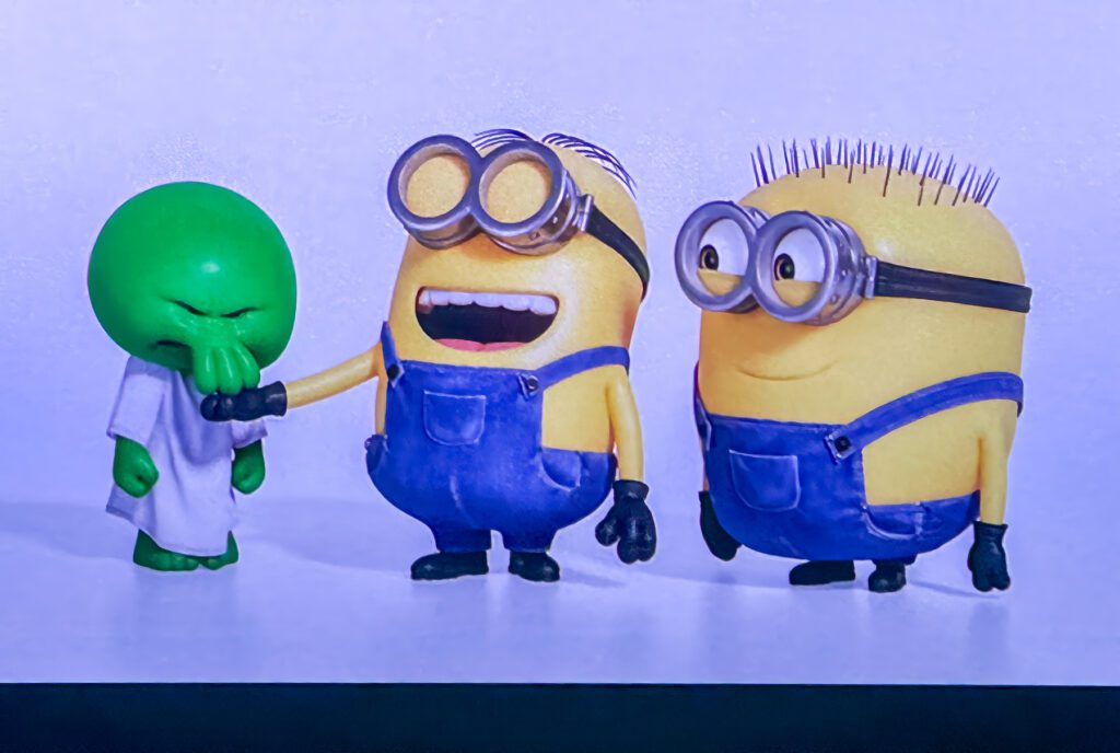 Two Minions in blue overalls stand next to a small green character in a white robe; one Minion is patting the green character’s head while smiling.