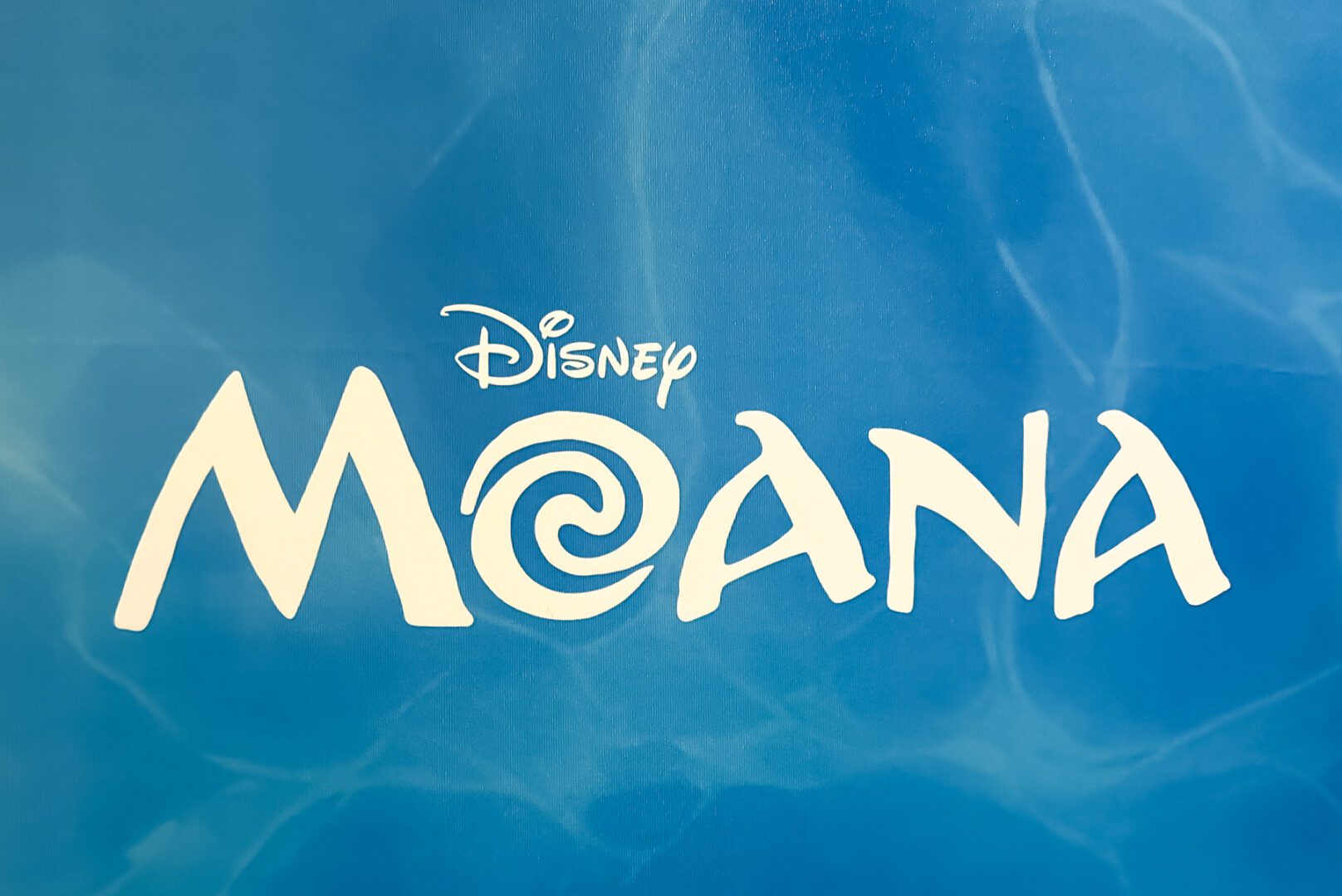 Disney "Moana" logo in white text with a spiral symbol, set against a blue, water-themed background.
