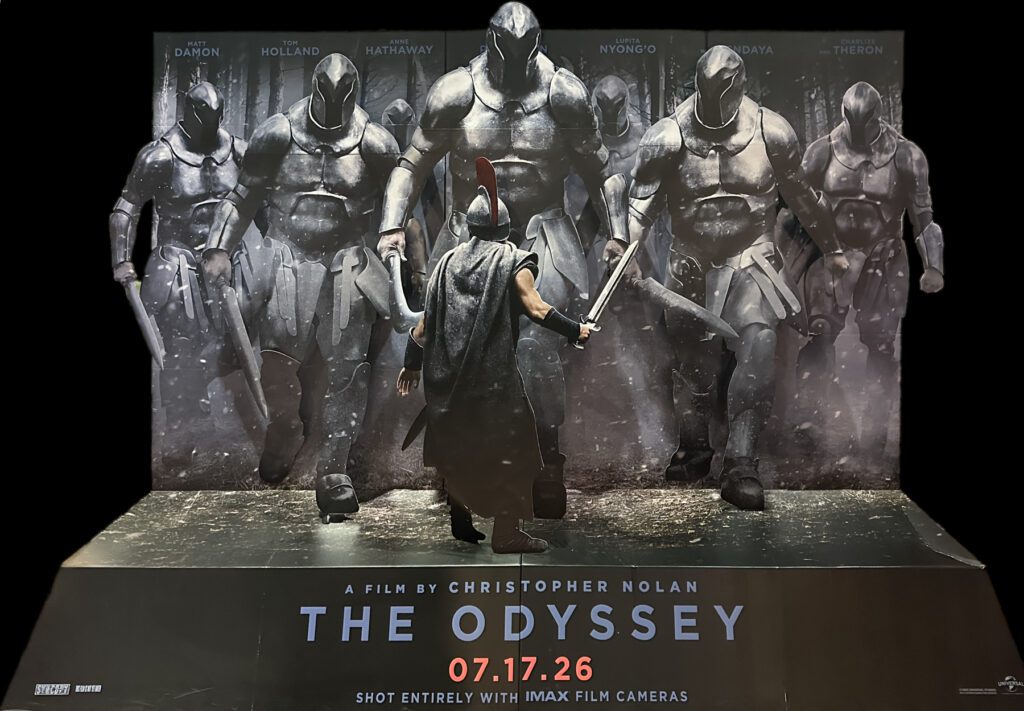A warrior in a helmet and cape faces six armored soldiers with swords in a promotional display for "The Odyssey" film by Christopher Nolan, releasing 07.17.26.