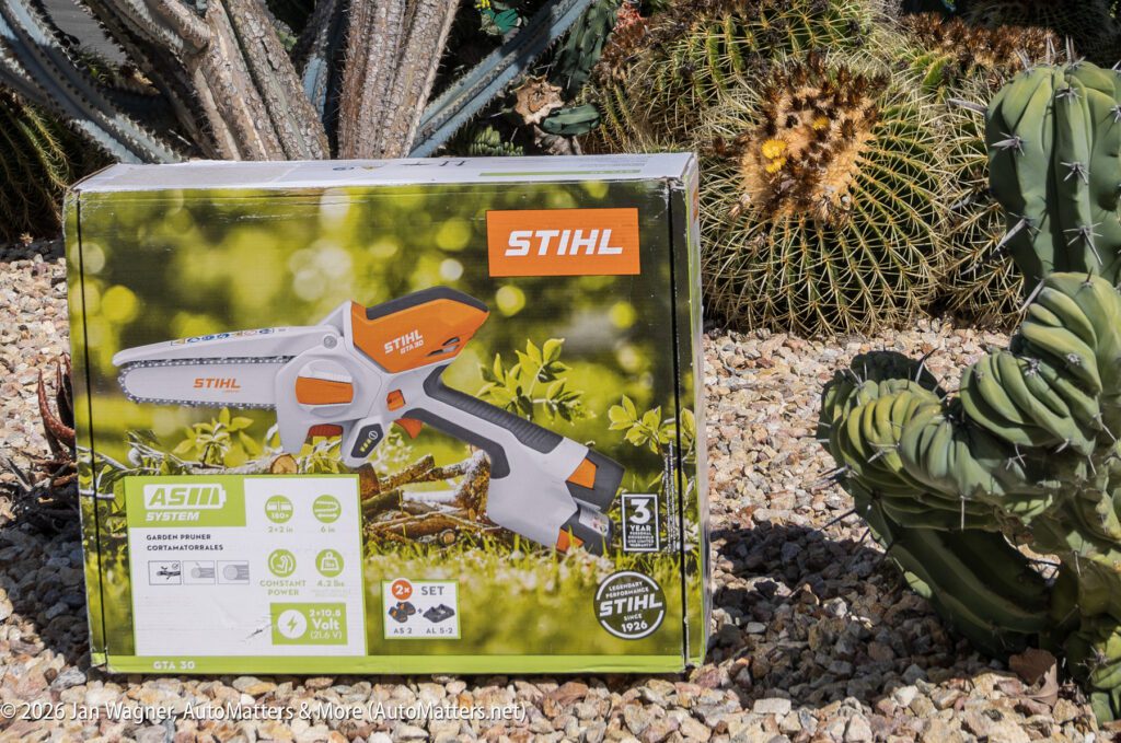 A Stihl GTA 26 cordless garden pruner box is displayed outdoors on gravel, surrounded by various cacti and succulents.