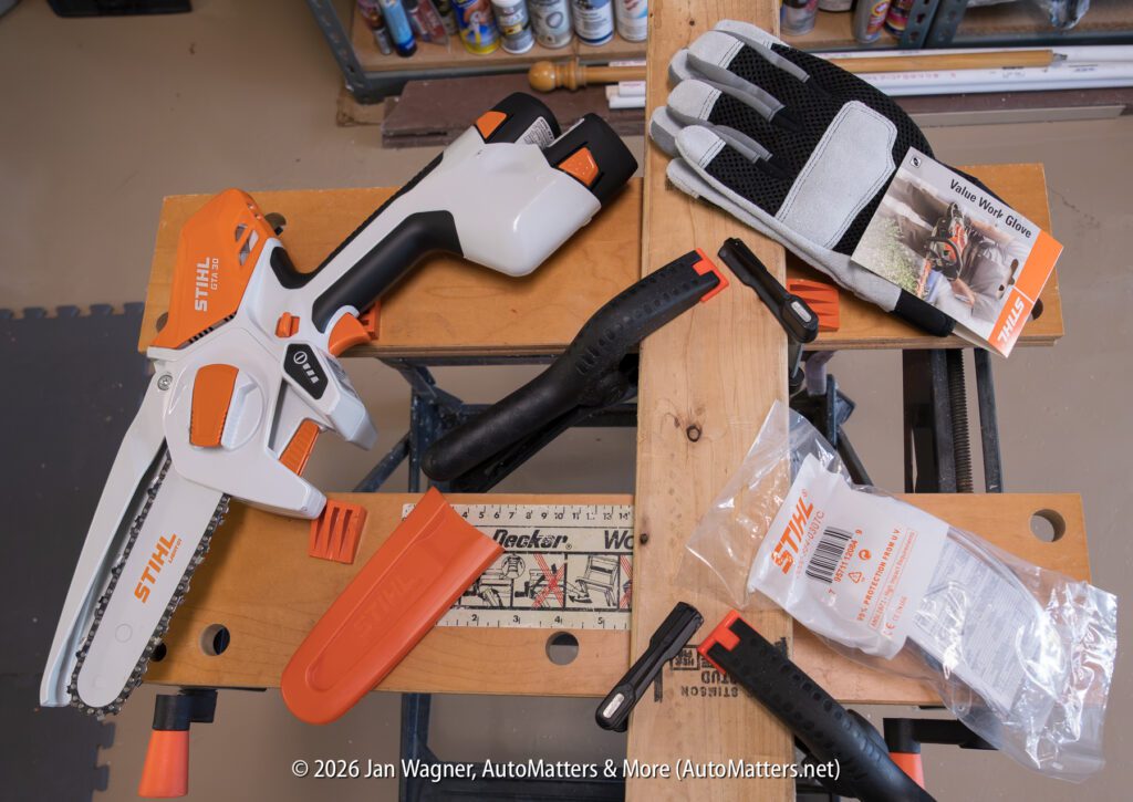 Stihl battery-powered mini chainsaw with gloves, manual, orange blade cover, clamp, wrench, and plastic bag on a workbench.