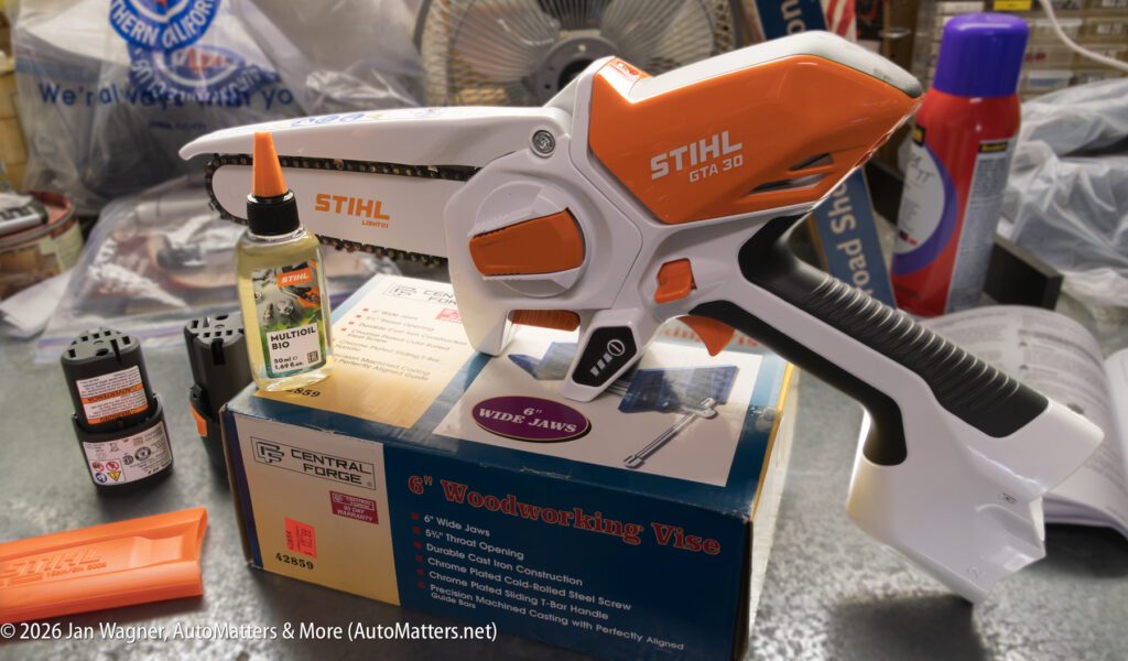 A Stihl GTA 26 cordless pruner is displayed on a box, with chain oil, accessories, and various workshop tools visible in the background.