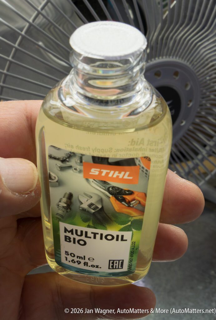 A hand holds a small 50 ml bottle of STIHL MultiOil Bio in front of a large metal fan.