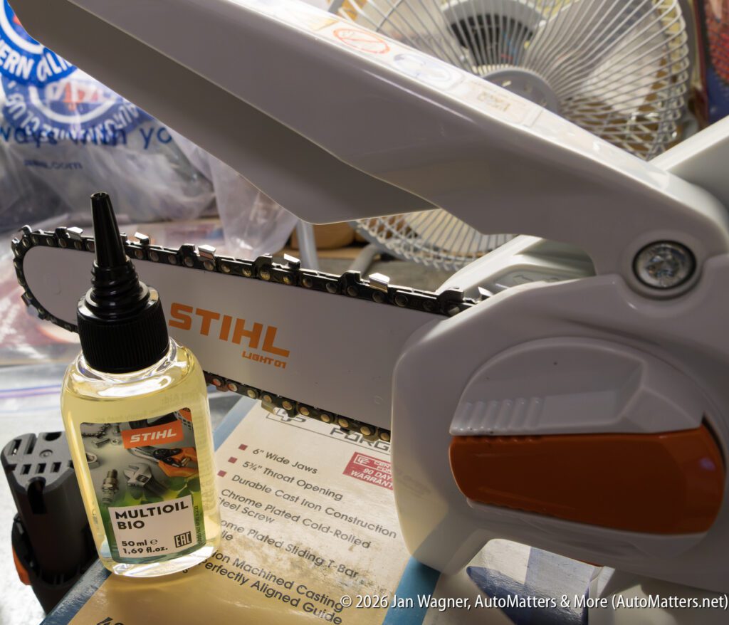 A Stihl electric chainsaw sits on a table next to a bottle of Stihl Multioil Bio lubricant, with a fan and assorted items in the background.