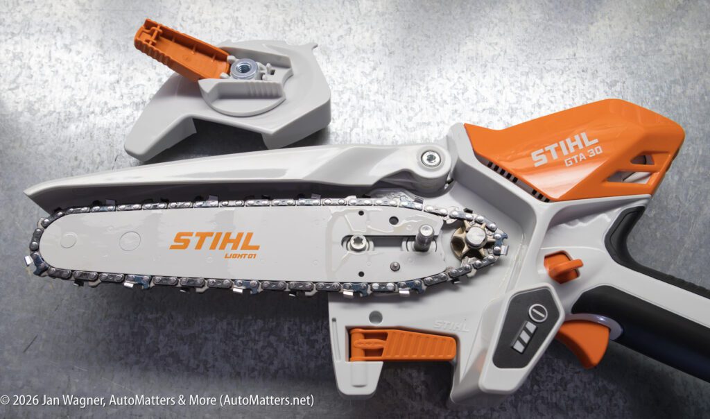 Close-up of a Stihl GTA 26 garden pruner with its orange and gray safety guard removed, showing the chain and cutting blade on a textured surface.