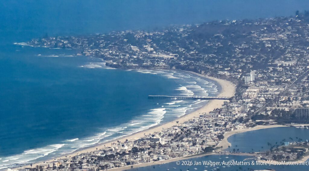 Aerial view of a coastal city with beaches, ocean waves, a pier, and densely packed buildings along the shoreline under a blue sky.