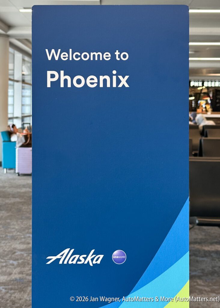 A blue sign at an airport reads “Welcome to Phoenix” with Alaska Airlines and oneworld logos. Passengers are seated in the background.