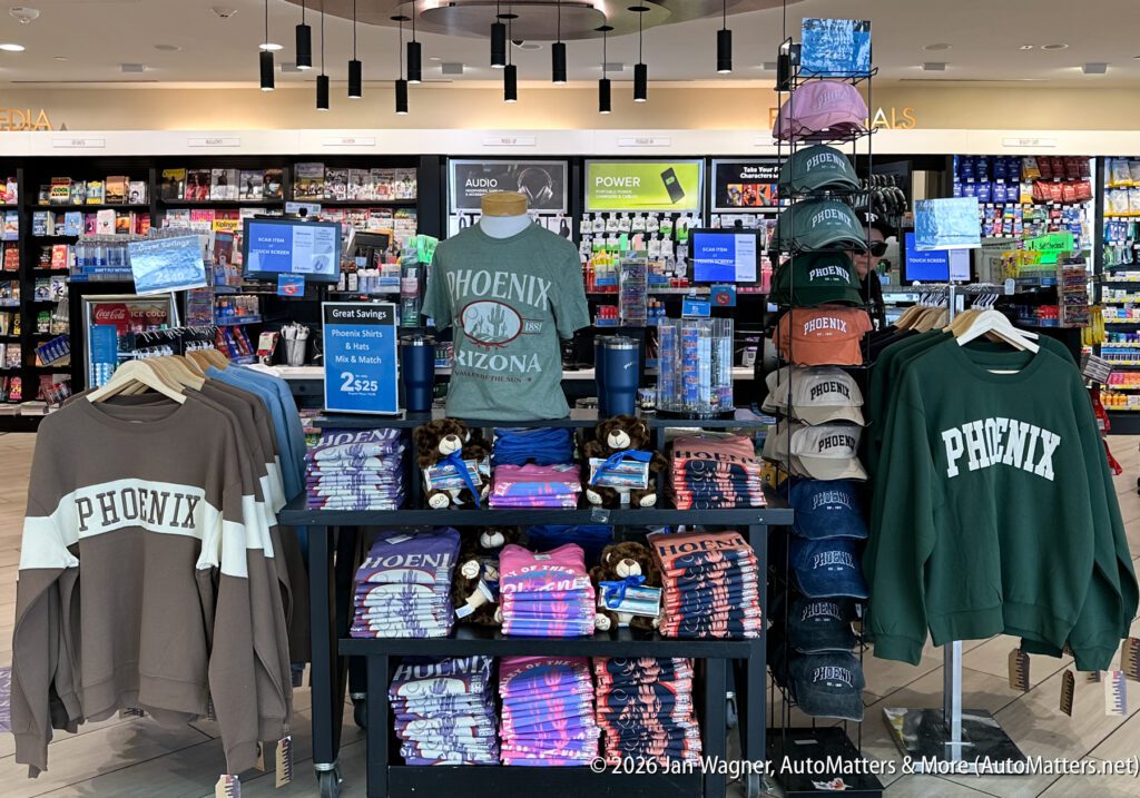 Display of Phoenix, Arizona-themed apparel, mugs, and souvenirs in an airport gift shop, with shirts, sweatshirts, and plush toys on tables and racks.
