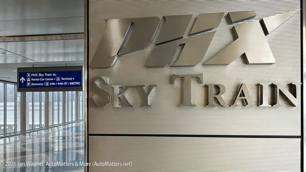 PHX Sky Train sign on a metal wall, with a blue overhead sign giving directions to rental cars, Terminal 4, economy parking, Valley Metro, and elevators.