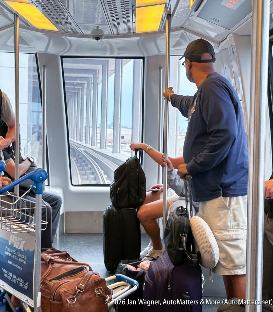 People sit and stand with luggage inside a train or tram, looking out large windows at railway tracks and a bridge structure ahead.