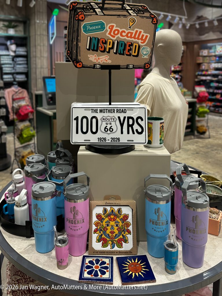A display of Phoenix-themed travel mugs, tumblers, decorative tiles, a Route 66 sign, and a “Locally Inspired” suitcase sign in a gift shop.