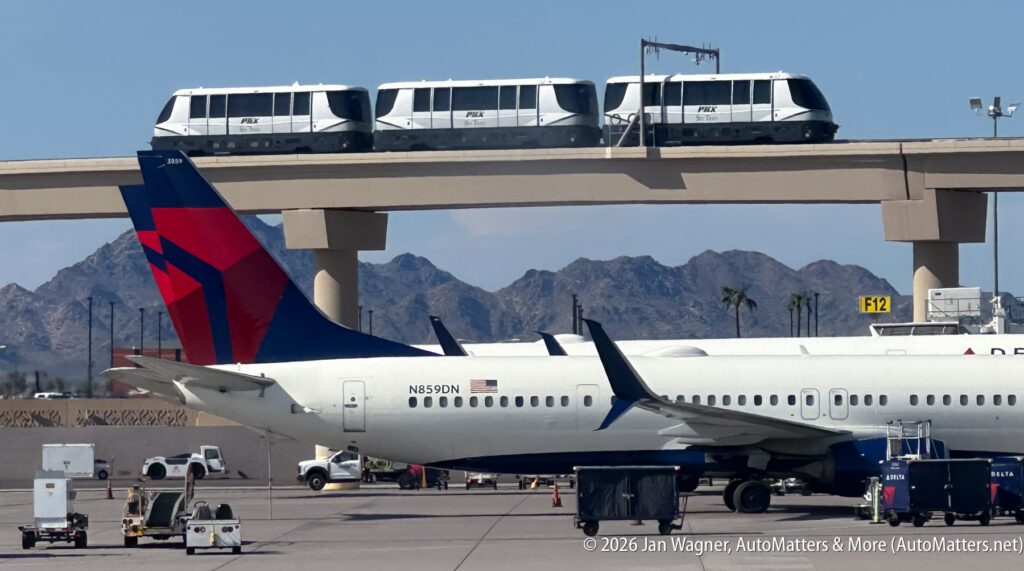 A Delta airplane sits at an airport gate with three PHX Sky Train cars traveling on an elevated track in the background.