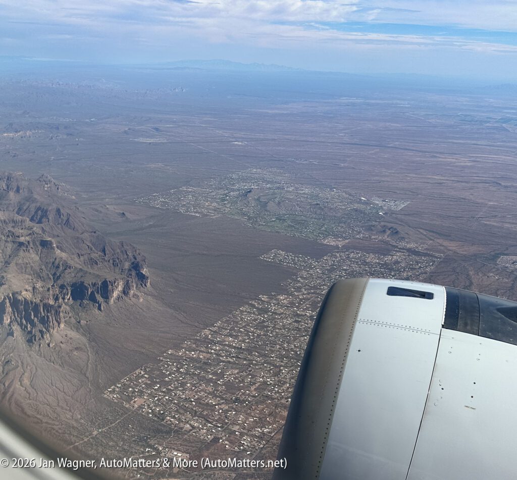 Aerial view of a desert landscape with scattered buildings and mountains, airplane engine visible in the foreground under a partly cloudy sky.
