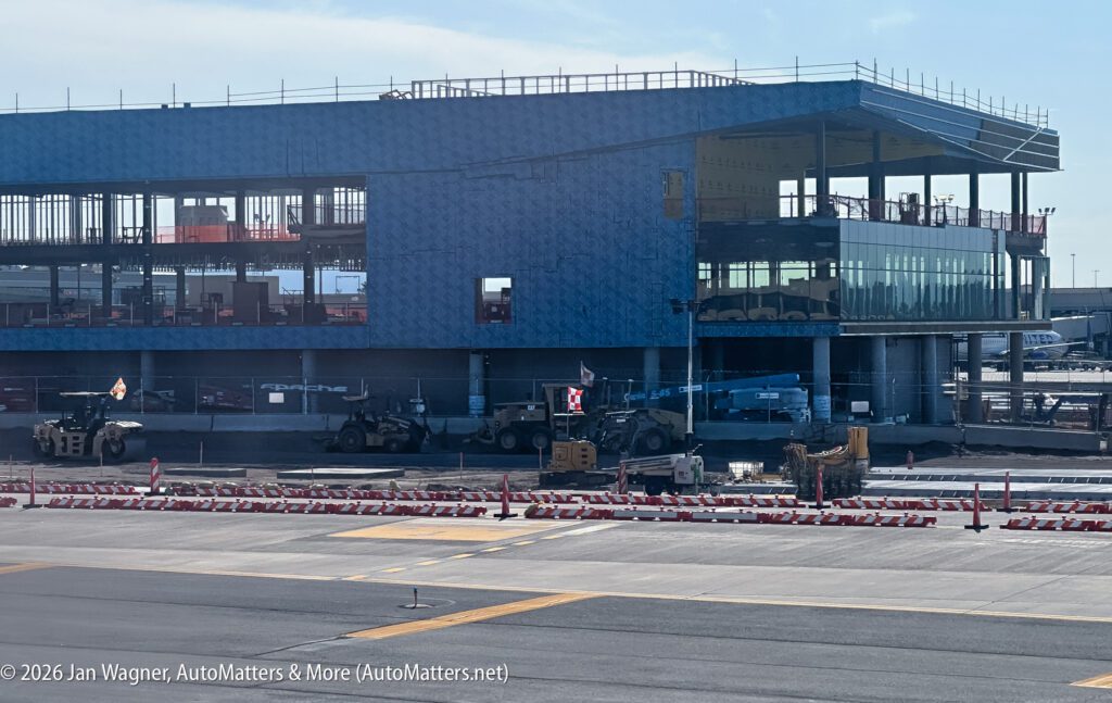Construction vehicles and equipment operate in front of a partially built, blue-wrapped airport terminal on a sunny day.