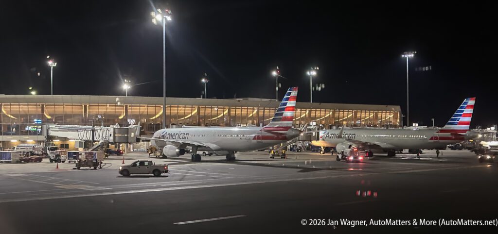 Two American Airlines planes are parked at gates outside a brightly lit airport terminal at night, with ground crew and vehicles nearby.